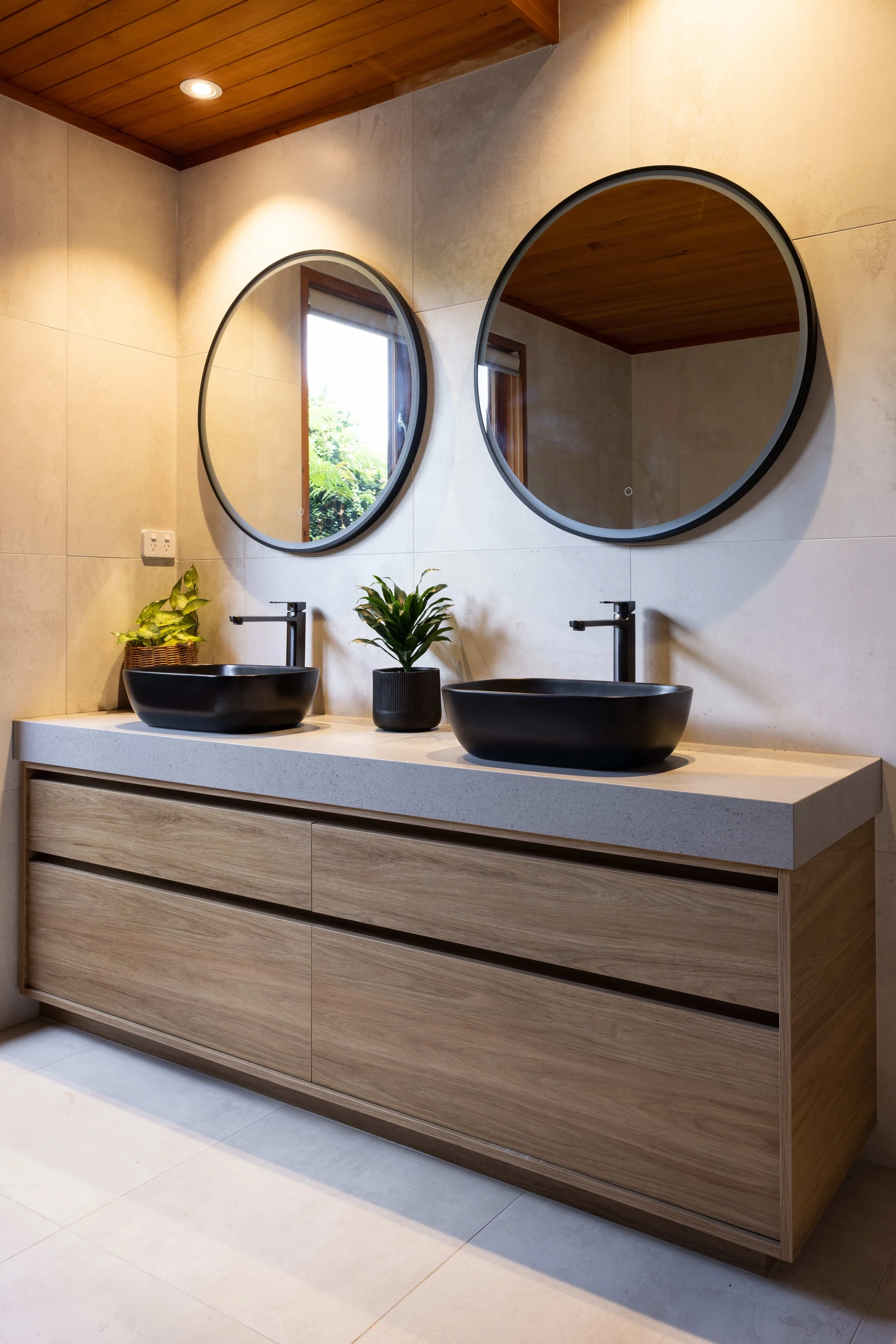 Modern bathroom vanity with two black vessel sinks, two round mirrors, a small potted plant, and a wooden cabinet beneath a beige countertop.