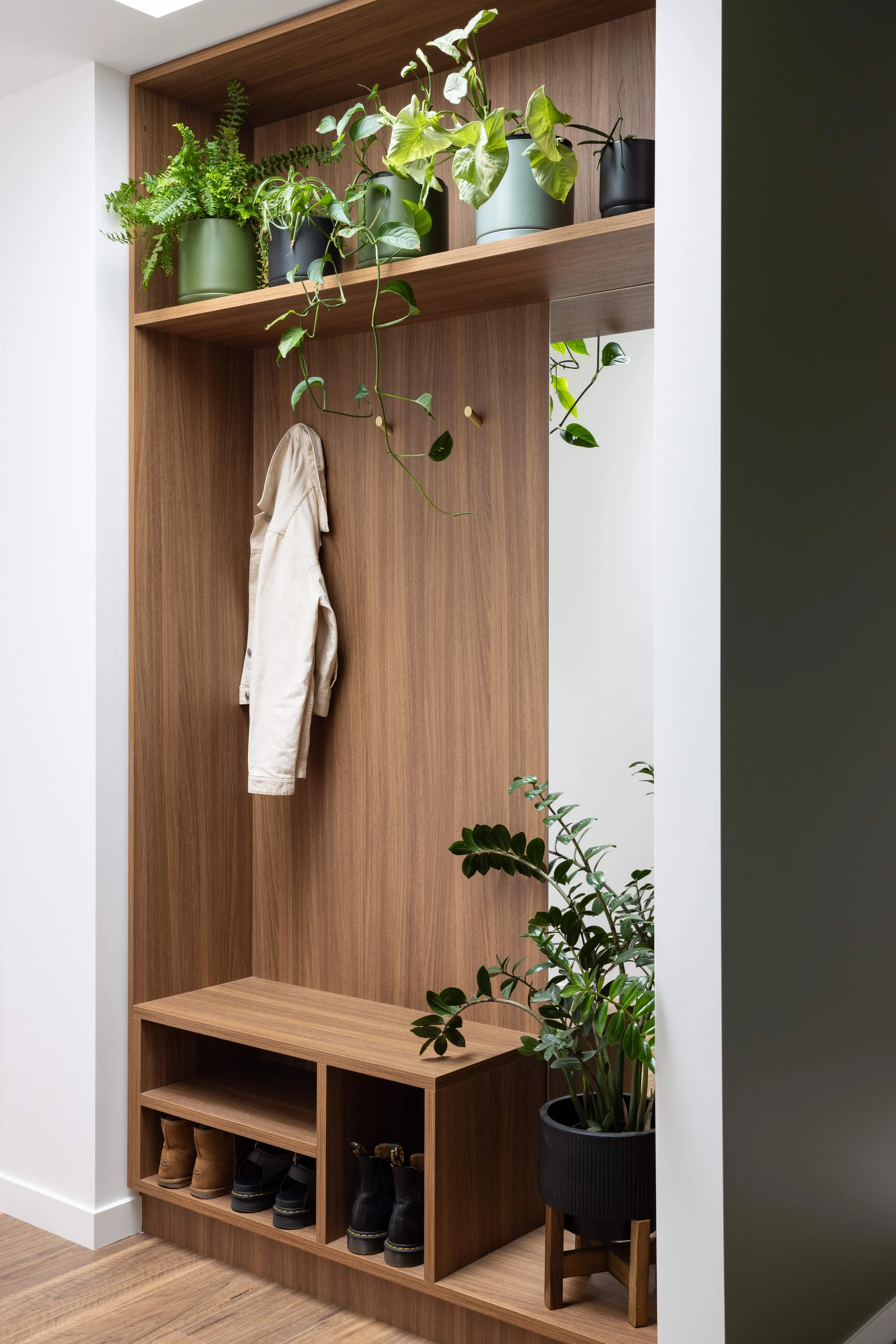 Modern entryway with wooden wall unit holding potted plants, a hanging coat, and shoes on a shelf.