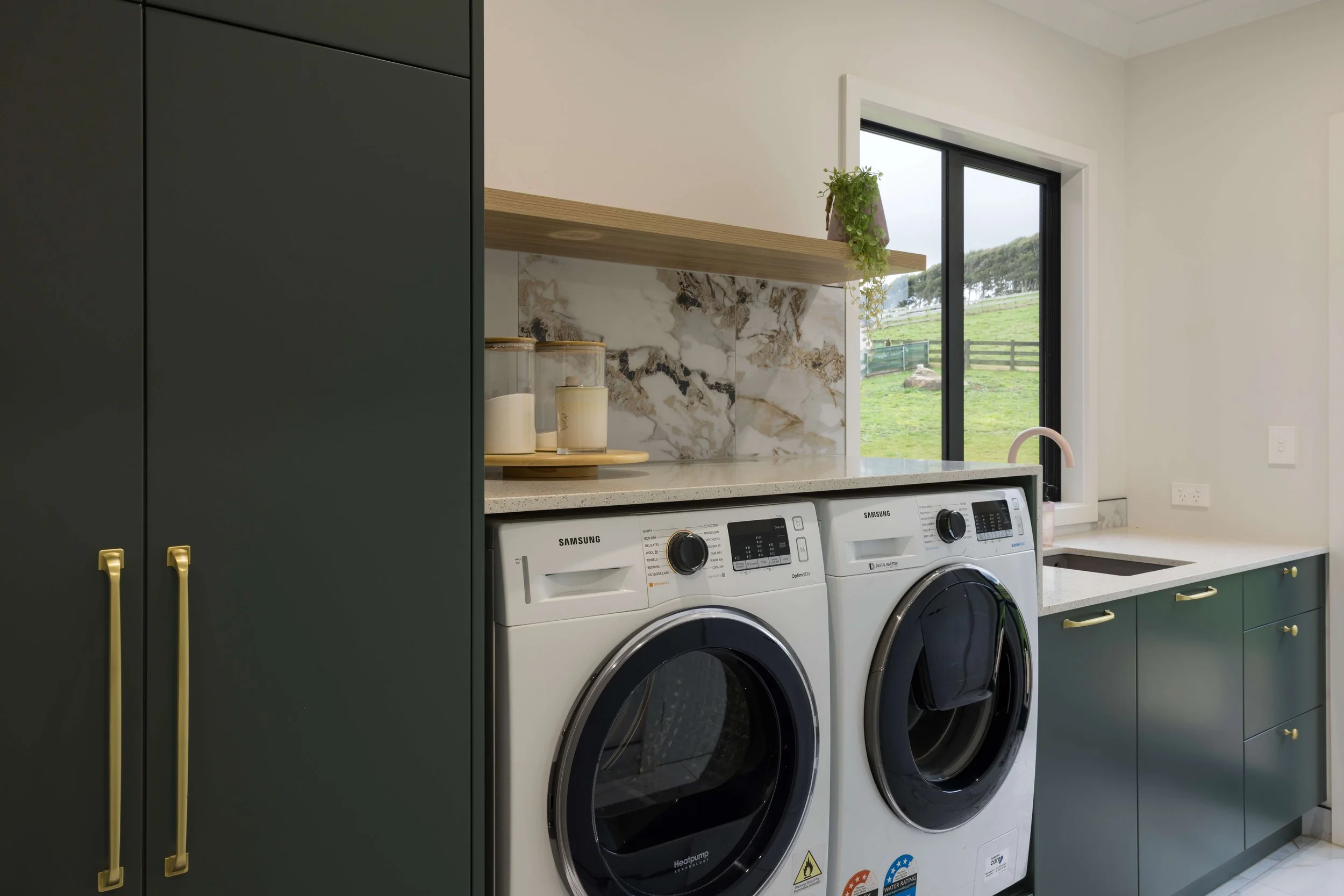 Laundry room with dark green cabinets, a white washing machine and dryer, a marble backsplash, a window showing a grassy outdoor landscape, and a small sink.