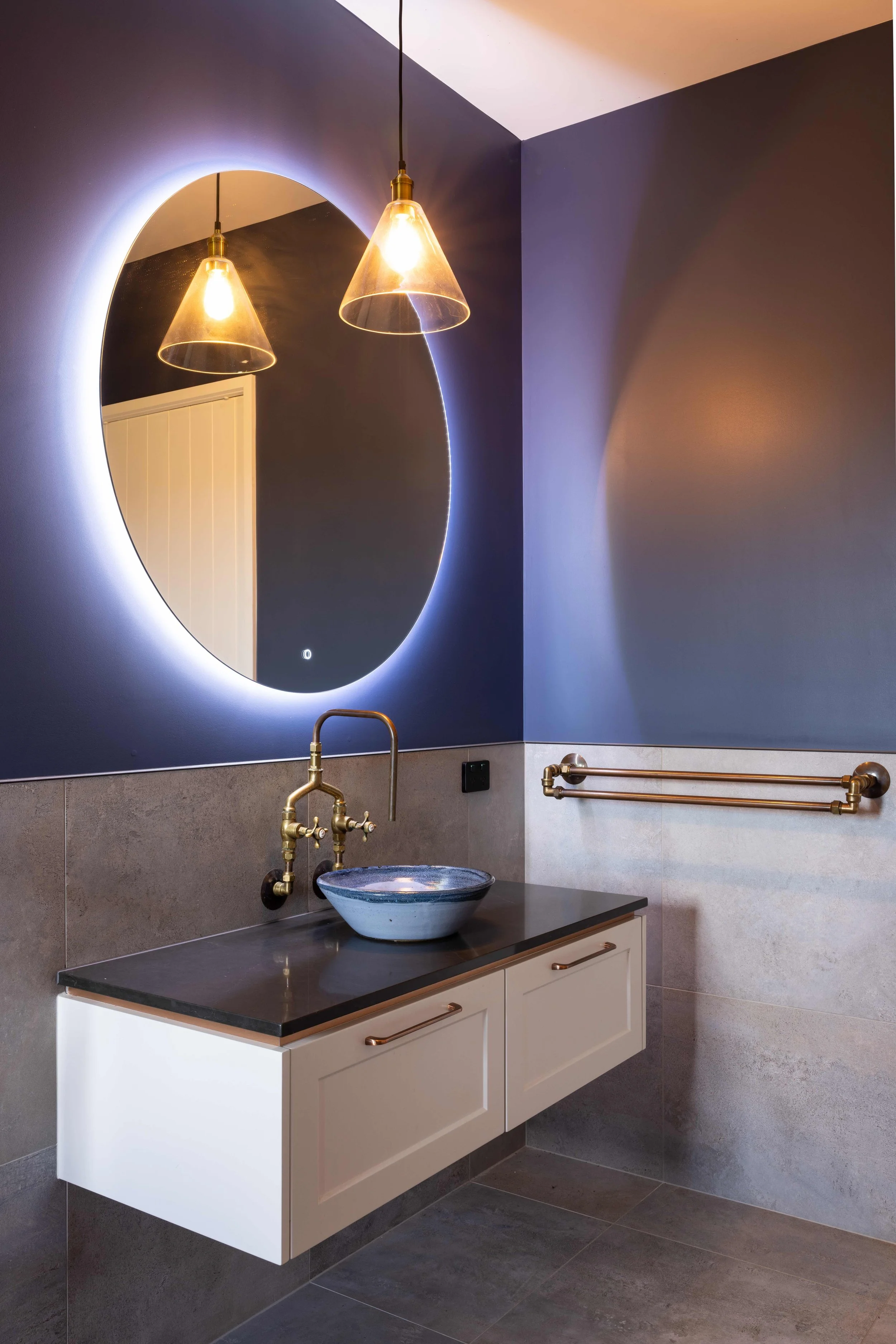A modern bathroom with a blue wall, circular mirror with backlighting, pendant lights, a vessel sink with brass fixtures, and a gold towel bar.