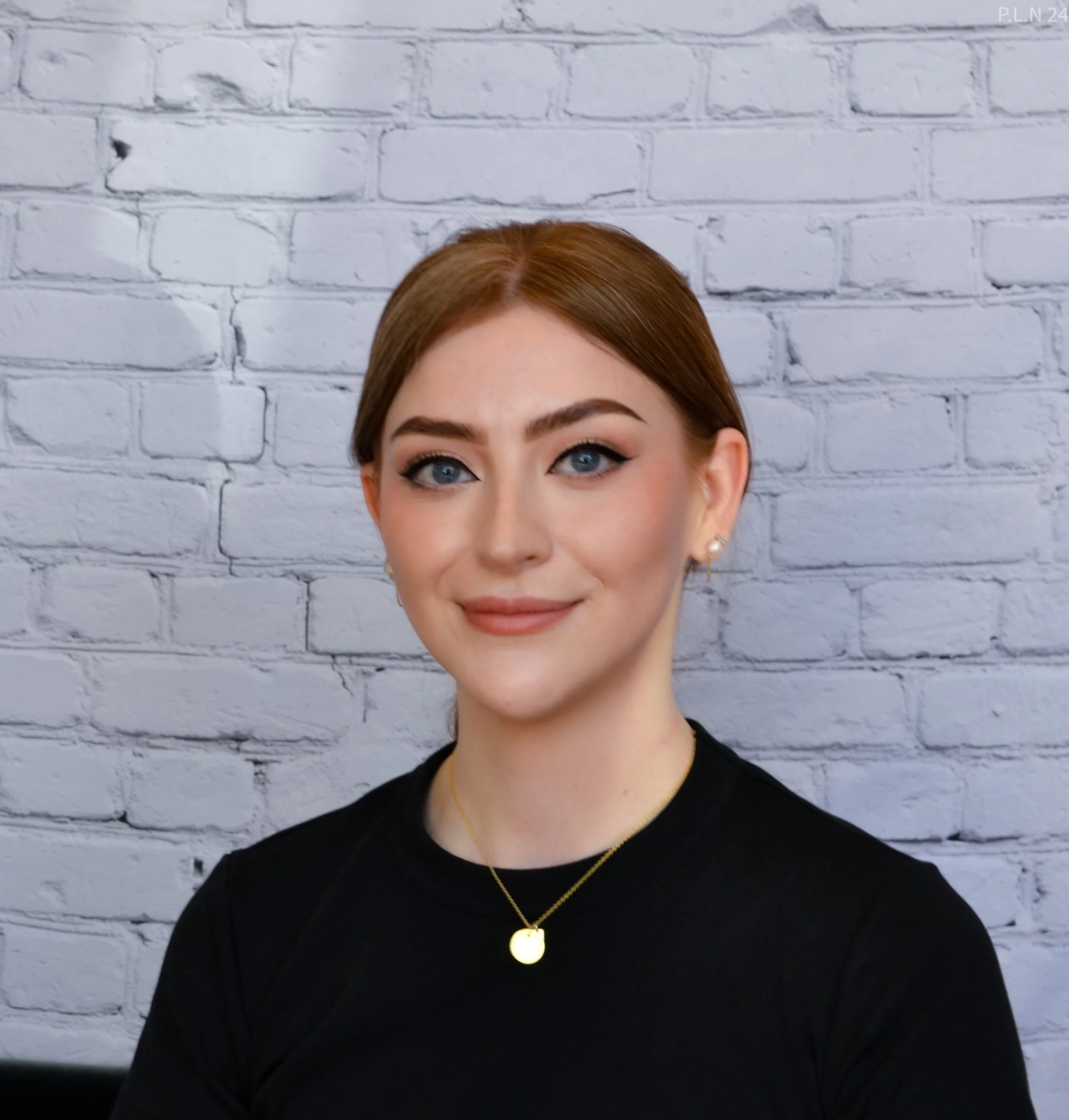 Portrait of a young woman with light skin, blue eyes, and brown hair styled back, wearing a black top and gold jewelry, standing against a white brick wall.