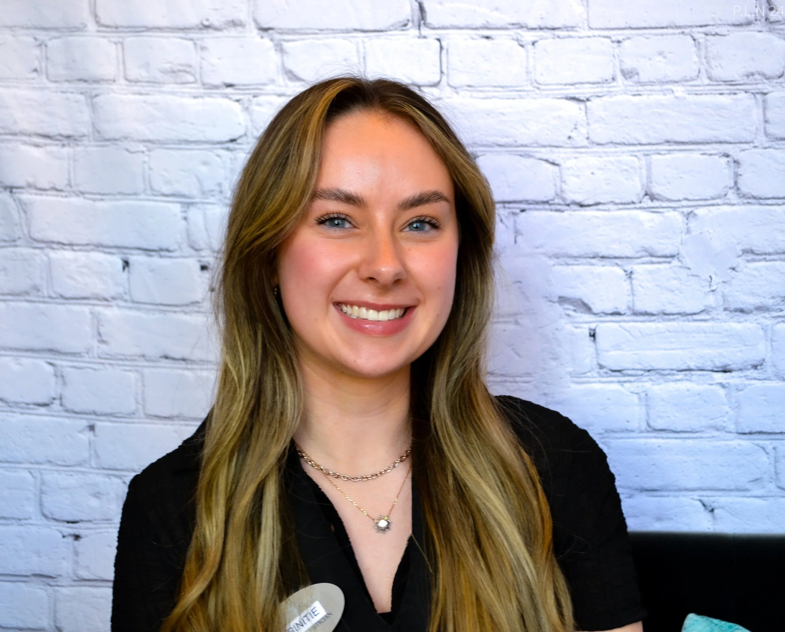A young woman with long blonde hair and blue eyes smiling, wearing a black top and layered necklaces, standing against a white brick wall.