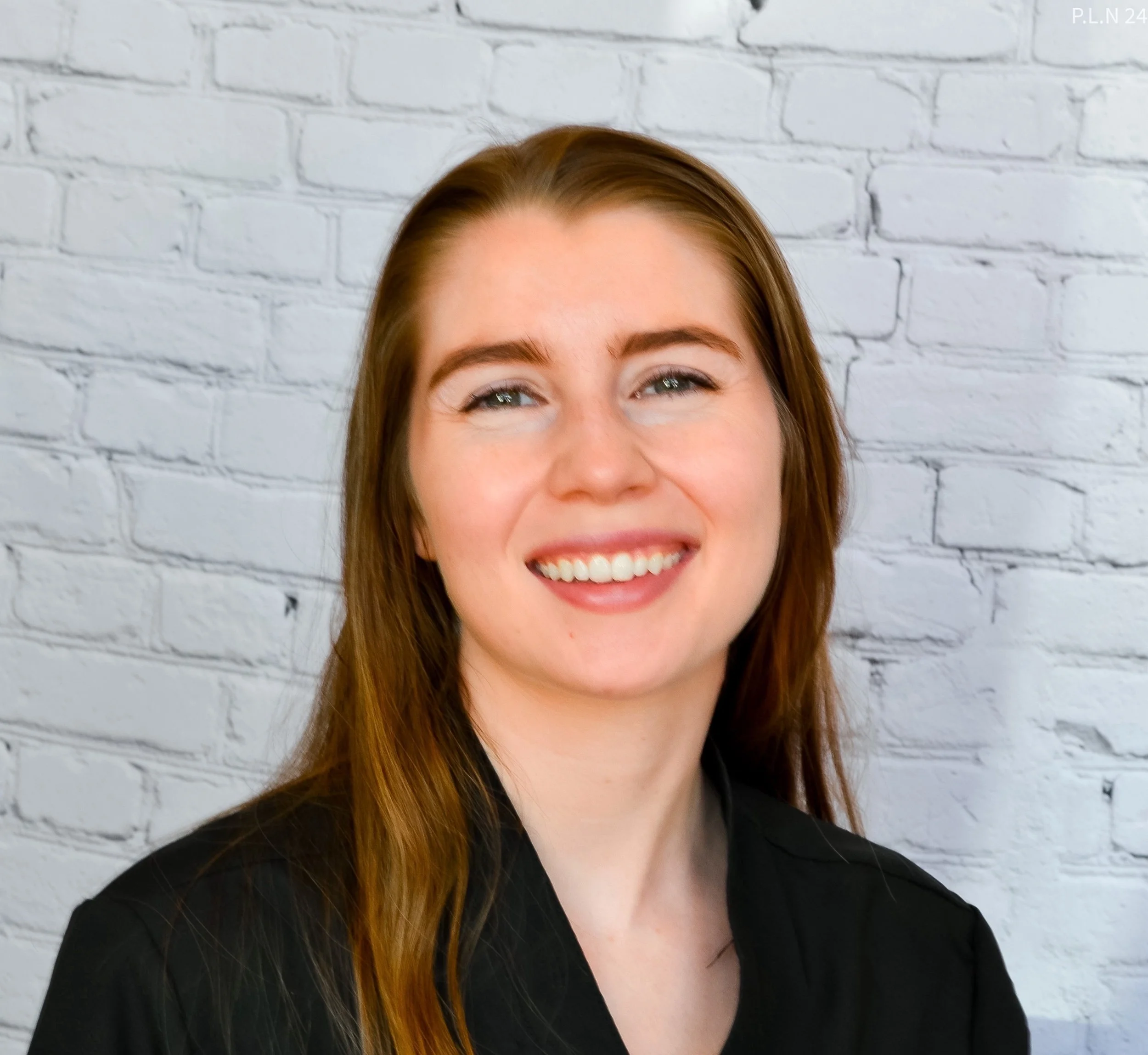 Smile of a woman with red hair and fair skin, wearing a black top, against a white brick wall background.