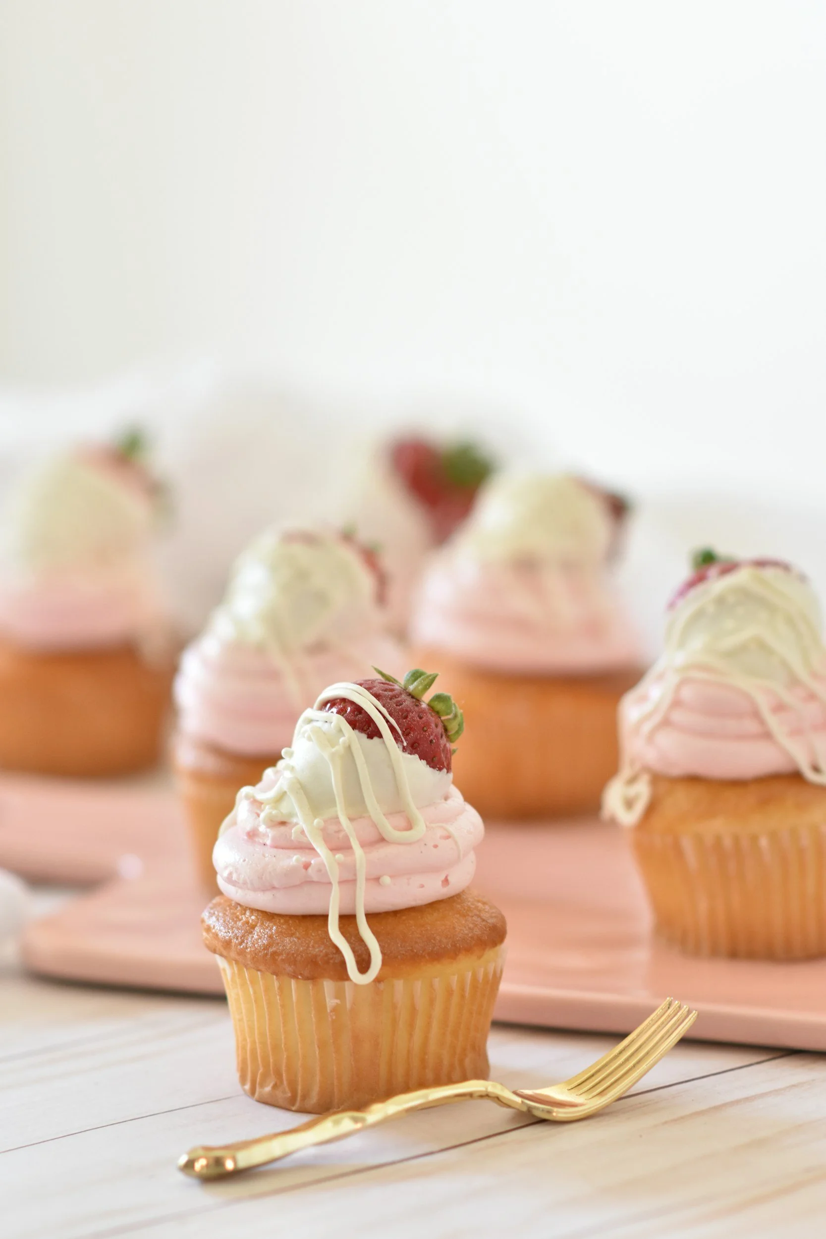 Cupcake with pink frosting, a strawberry on top, and white chocolate drizzled over it, placed on a white surface with a golden fork beside it.