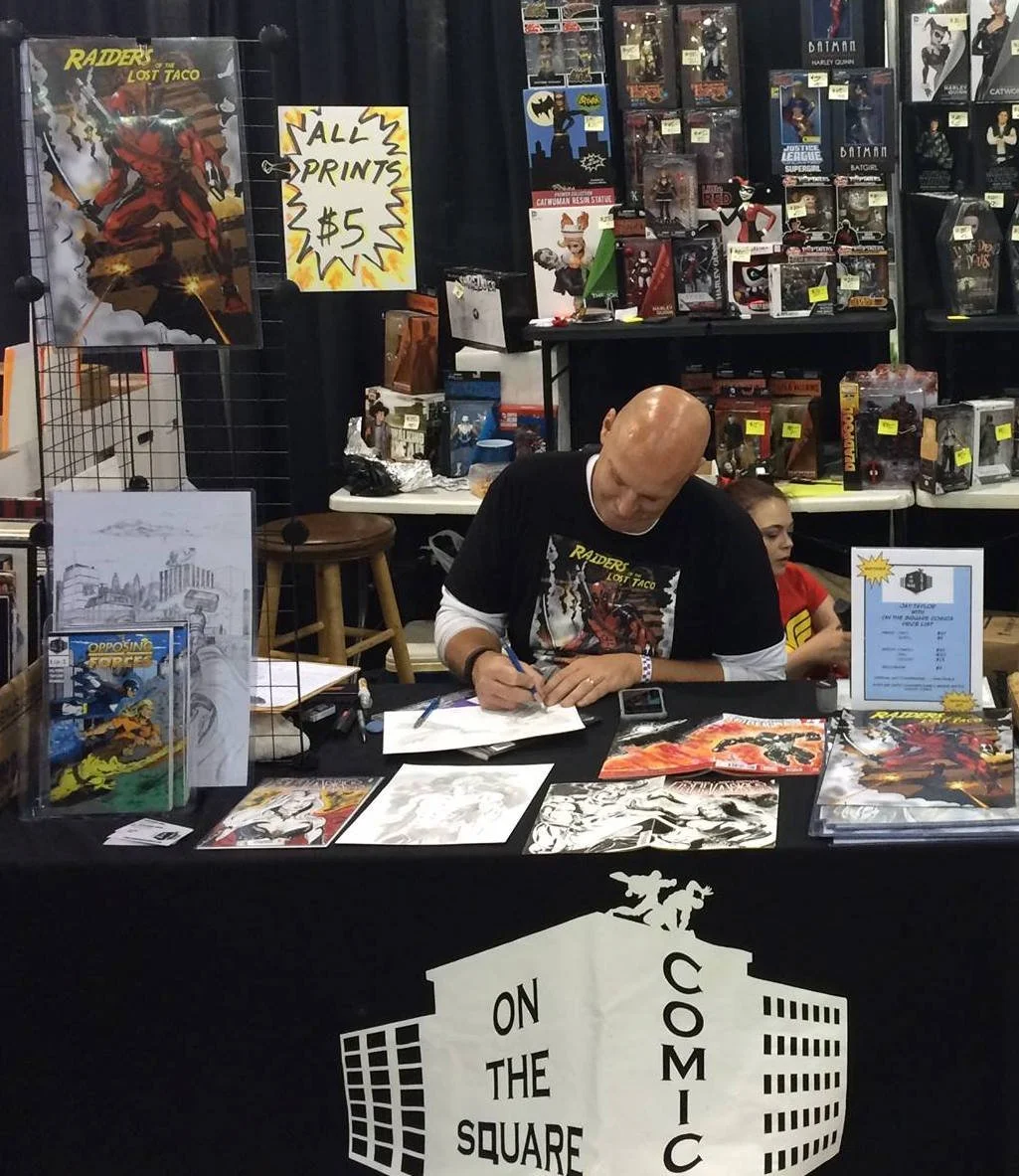 Jay Taylor signing artwork at a convention booth, surrounded by comic books and memorabilia. The booth has a sign that reads 'On the Square Comic'