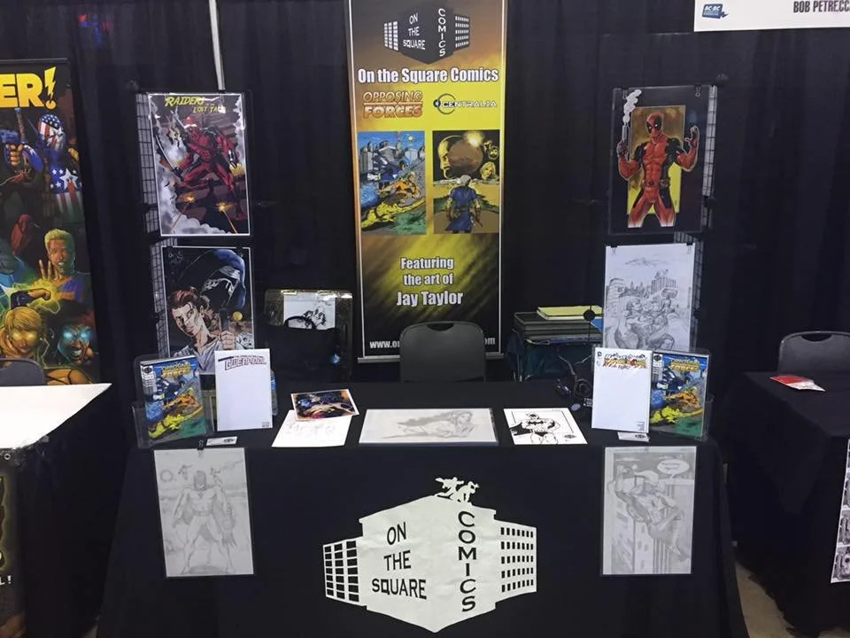 Booth at a comic convention featuring display banners, artwork, comic books, and promotional materials for On the Square Comics, with a black tablecloth bearing the company's logo and a black backdrop.