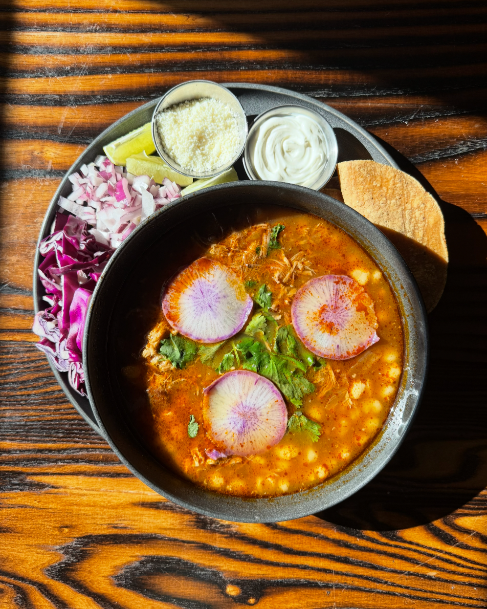 A bowl of soup on a plate with tortillas, crema, cotija, radishes, and lime wedges on a wooden tabletop.