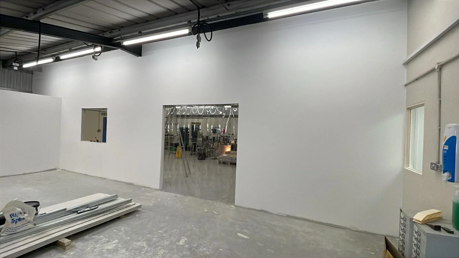 Interior of a building under construction with white walls, a partial wall with a rectangular opening, construction materials on the floor, and a glimpse into a room with furniture.
