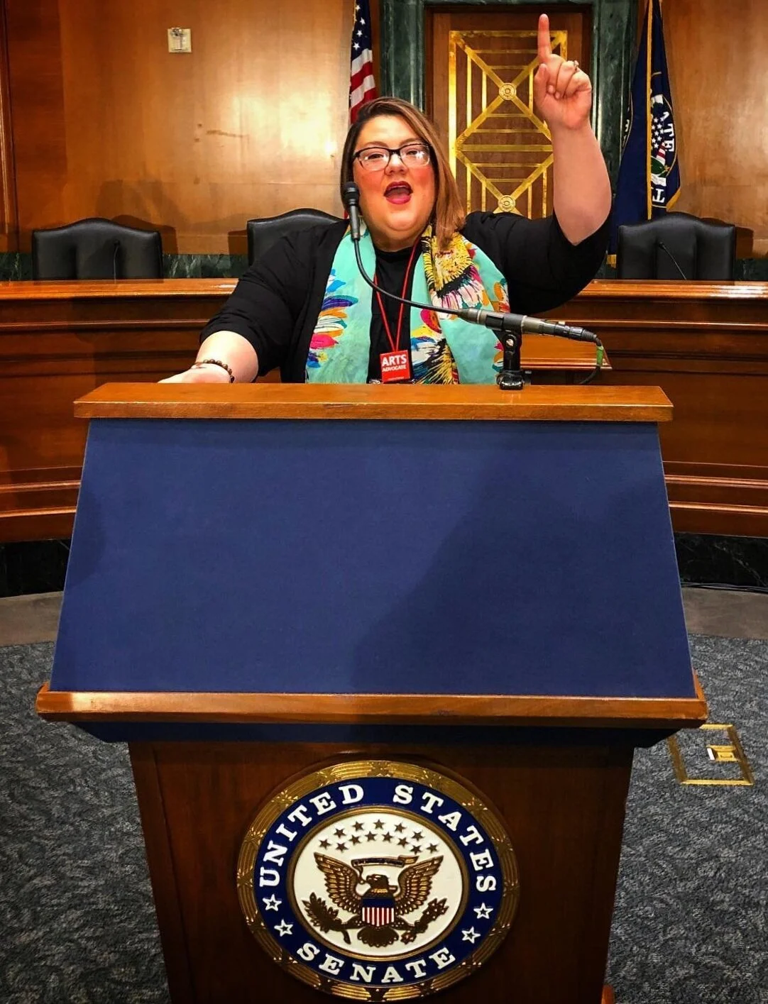 A woman with glasses and shoulder-length hair, wearing a colorful scarf and a black outfit, is speaking at a U.S. Senate podium with a microphone, raising her right hand with her index finger pointing upward. The background features wooden paneling, flags, and the official U.S. Senate seal.
