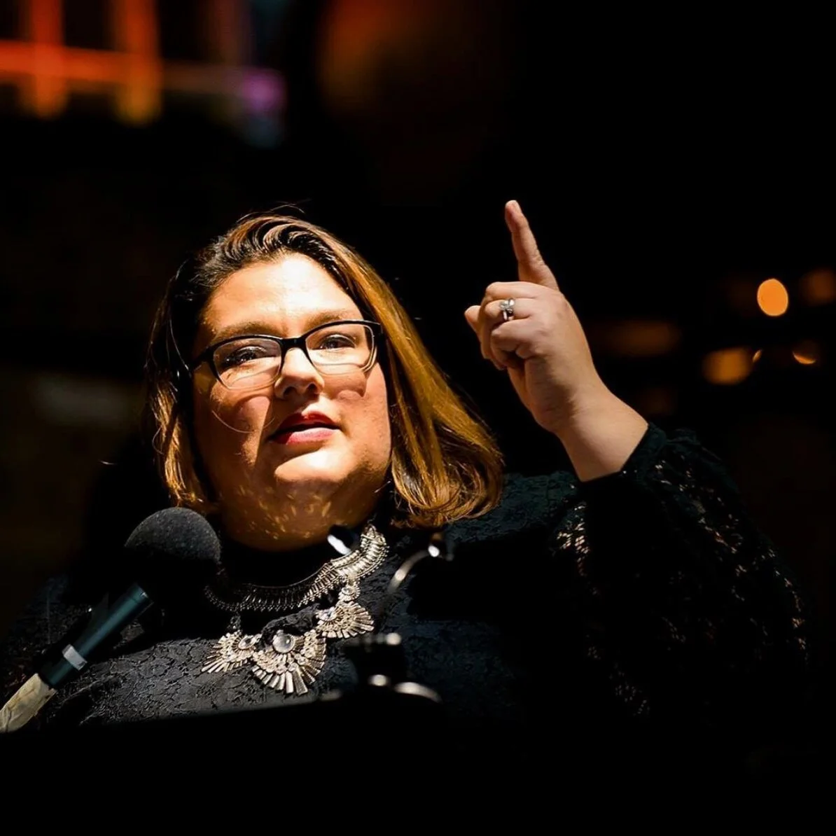 A woman with glasses and wearing a black lace top, speaking into a microphone with a serious expression, pointing upward with her right index finger.