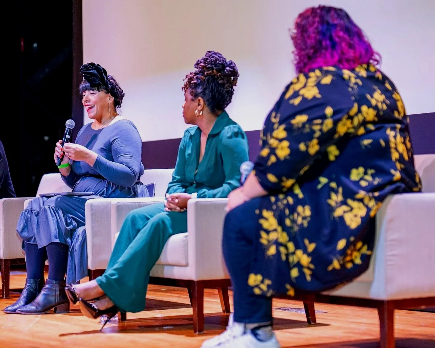 Three women sitting on chairs on a stage during a panel discussion. One woman is speaking into a microphone, while the other two women listen.