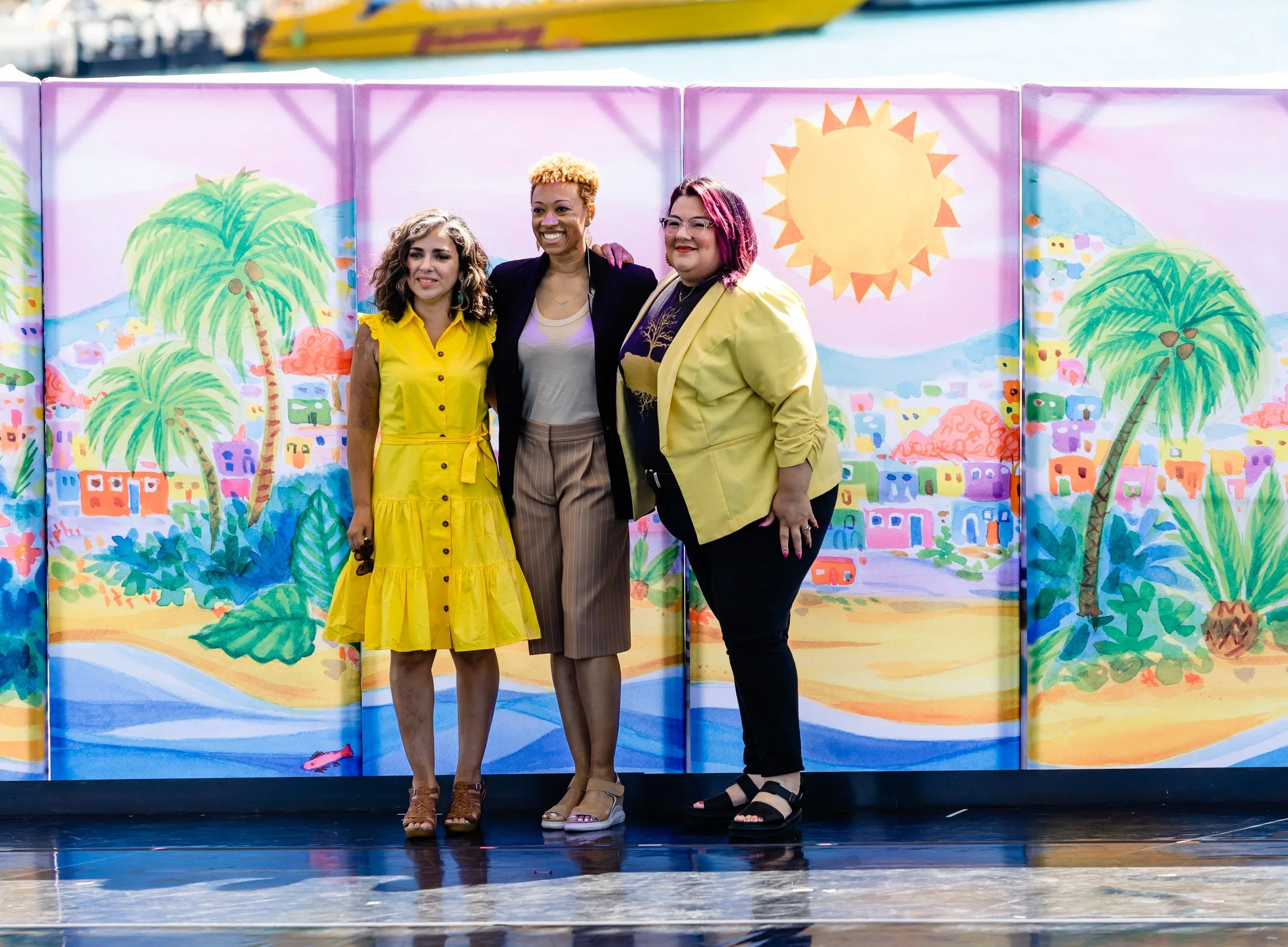 Three women standing together on stage in front of a colorful painted backdrop of a tropical scene with palm trees, hills, and a bright sun.
