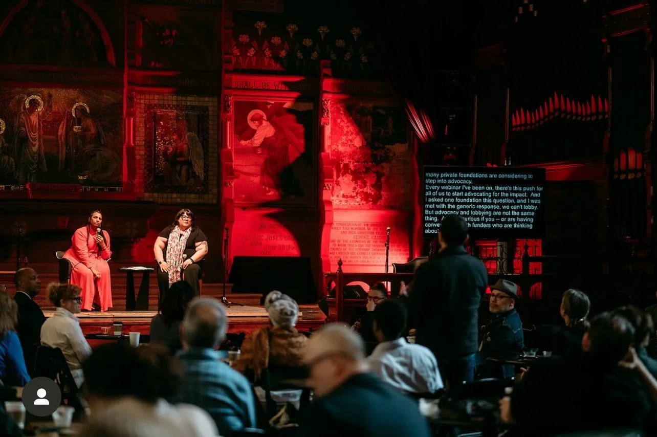 Two women are sitting on stage during a discussion at a formal event, with an audience watching. One woman is wearing a red dress, and the other is wearing a black top with a scarf. The stage background is decorated with large historical or religious paintings and a red lighting effect.