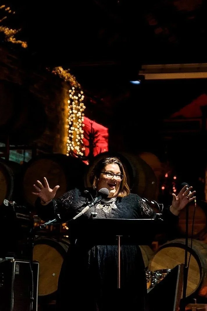 A woman with glasses, dark hair, and a black lace dress speaking at a podium with a microphone, with Christmas lights and wooden barrels in the background.