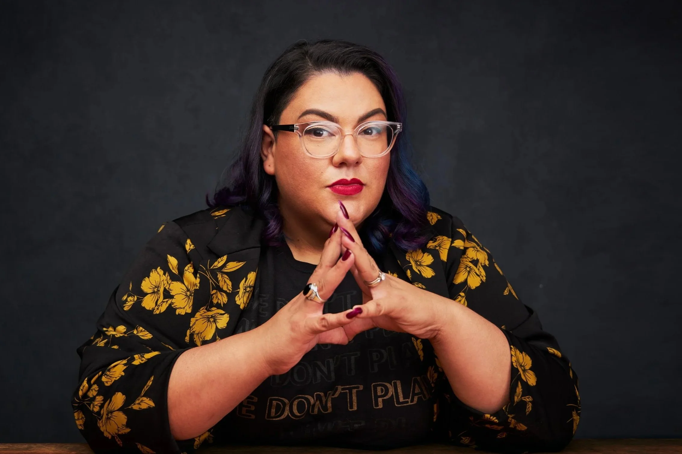 A woman with dark hair with purple highlights, wearing glasses, a black shirt with partly visible text, and a black jacket with yellow floral pattern, sitting against a dark background with her hands clasped in front of her face.