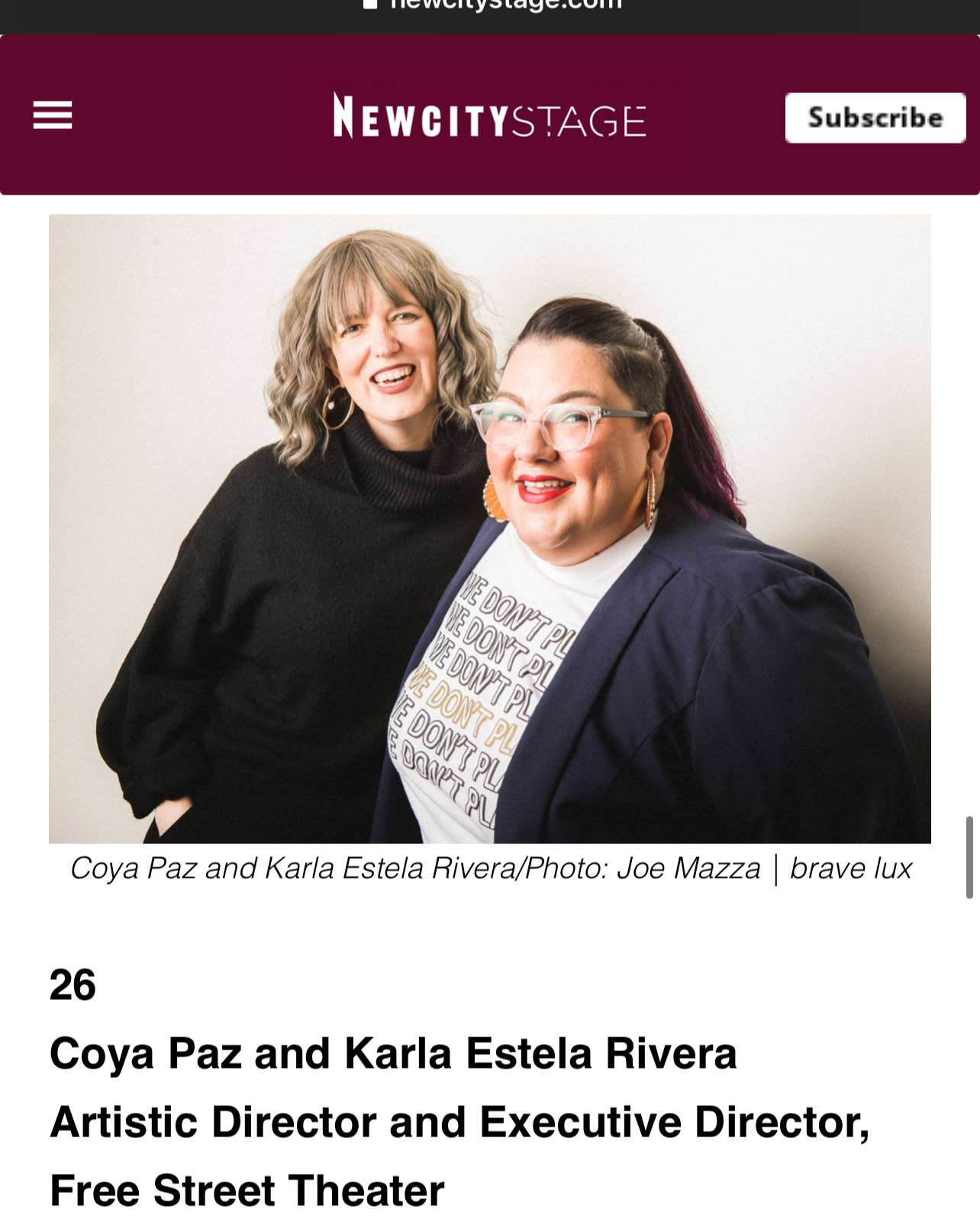 Two women smiling and posing for a photo in front of a plain wall. One woman has curly, shoulder-length hair, wears a black turtleneck, and has gold hoop earrings. The other woman has short, dark hair with purple tips, wears glasses, a white shirt with text, a navy blazer, and large gold earrings.