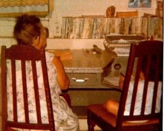 A young girl sitting at a desk with books and papers, facing away from the camera, in a room with a bookshelf and window in the background.