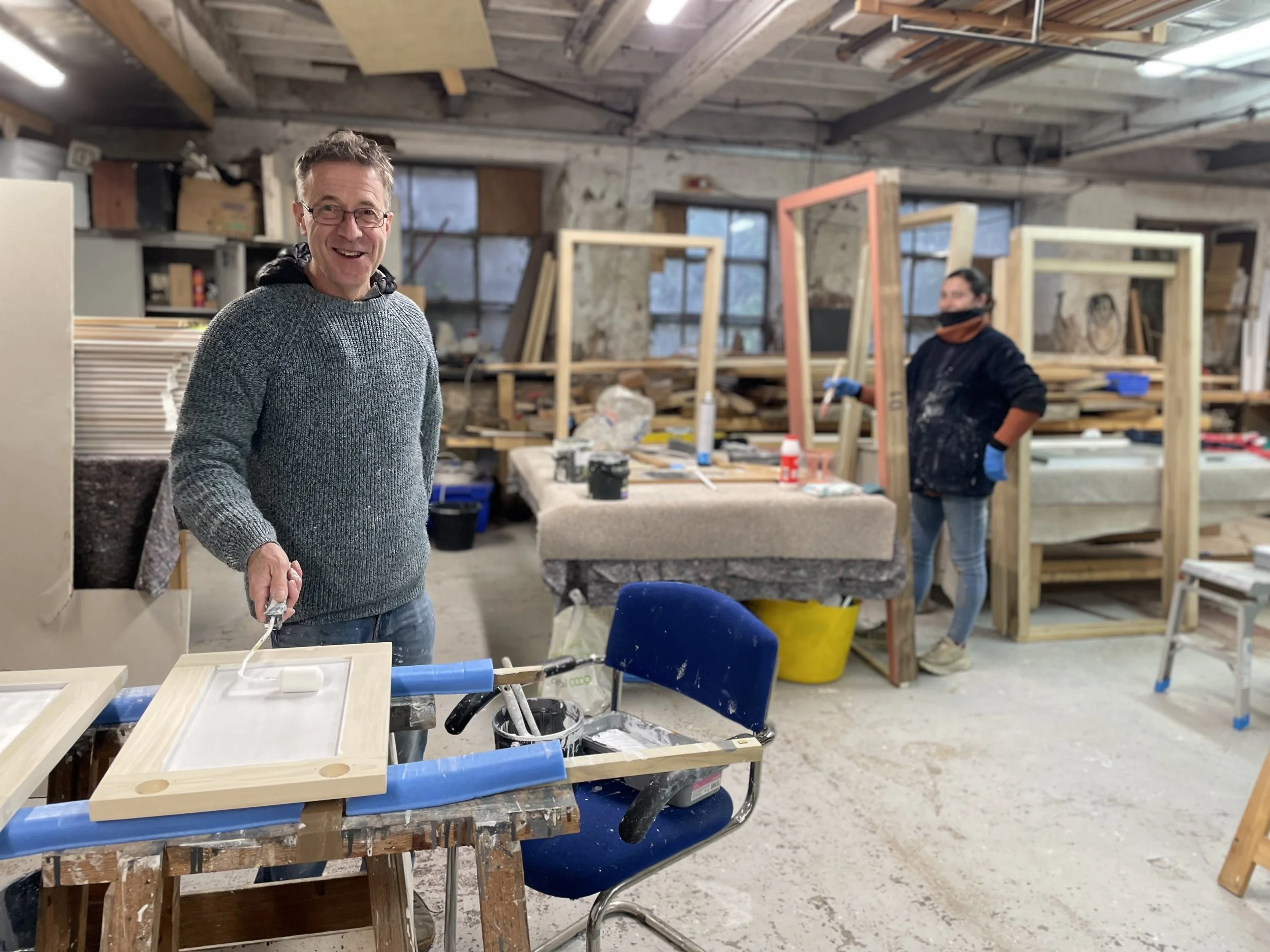 Pete the painter smiles at the camera as he paints a window frame in the Water lane joinery workshop