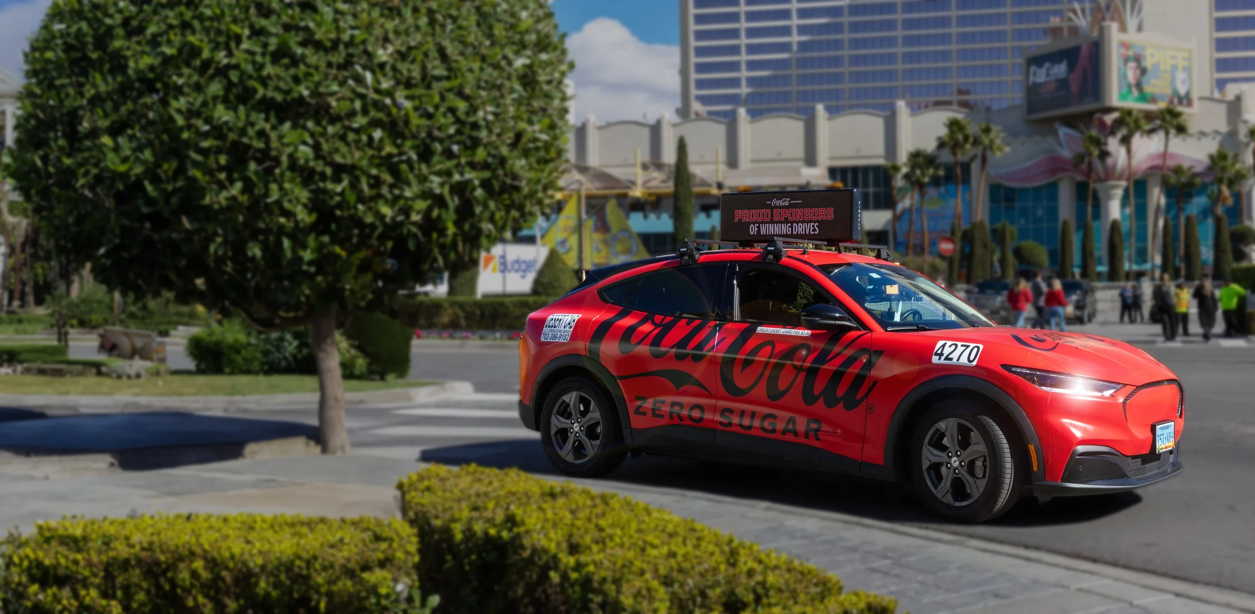 Red car with Coca-Cola branding parked on city street, surrounded by trees and buildings, with people in the background.