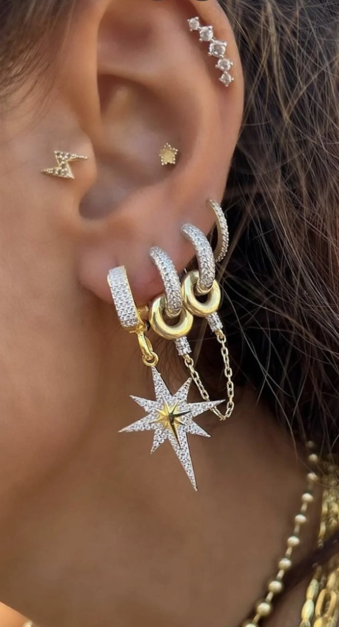Close-up of a person's ear adorned with multiple earrings, including a star-shaped stud, a lightning bolt stud, a row of small diamonds along the helix, and several hoop earrings with dangling star and chain charms.