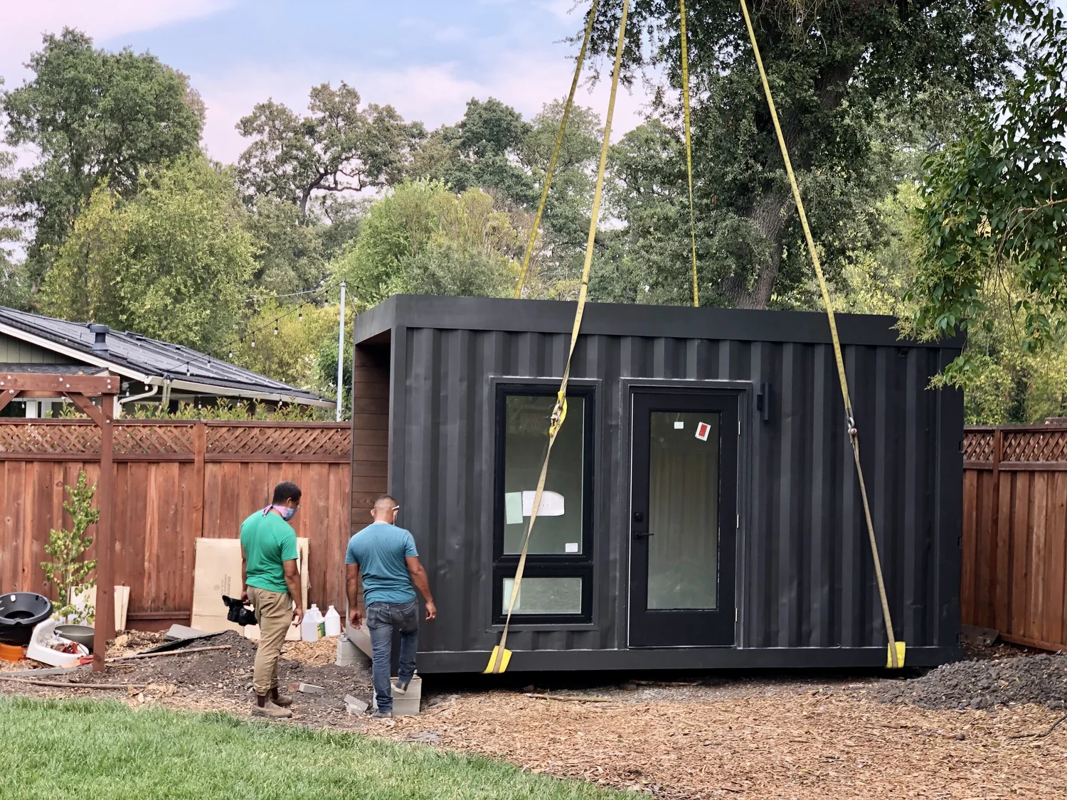 Two installers guide a dark ADU module as a crane lowers it into a fenced backyard