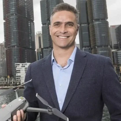 Man smiling in business attire holding a drone with city skyscrapers in the background.