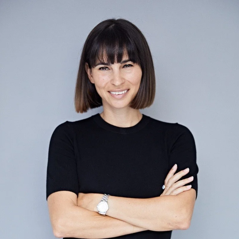 A woman with shoulder-length dark hair and bangs, smiling with arms crossed, wearing a black short-sleeve top and a watch, standing against a plain gray background.