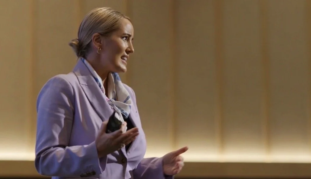 A woman with blonde hair in a bun, wearing a lavender blazer and blouse, standing and speaking in front of a warm-colored background.