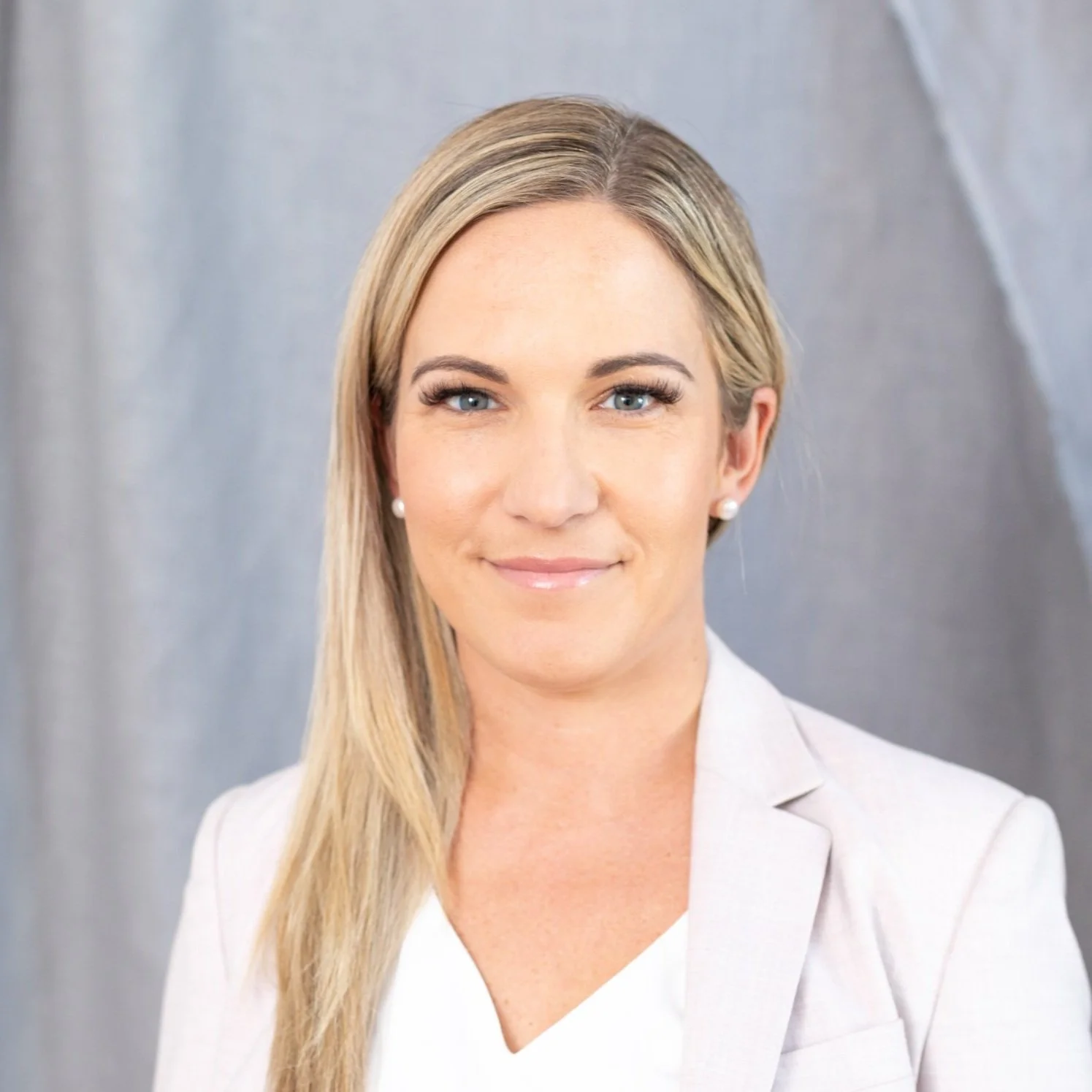 Portrait of a woman with blonde hair, blue eyes, wearing pearl earrings and a light-colored blazer against a gray background.