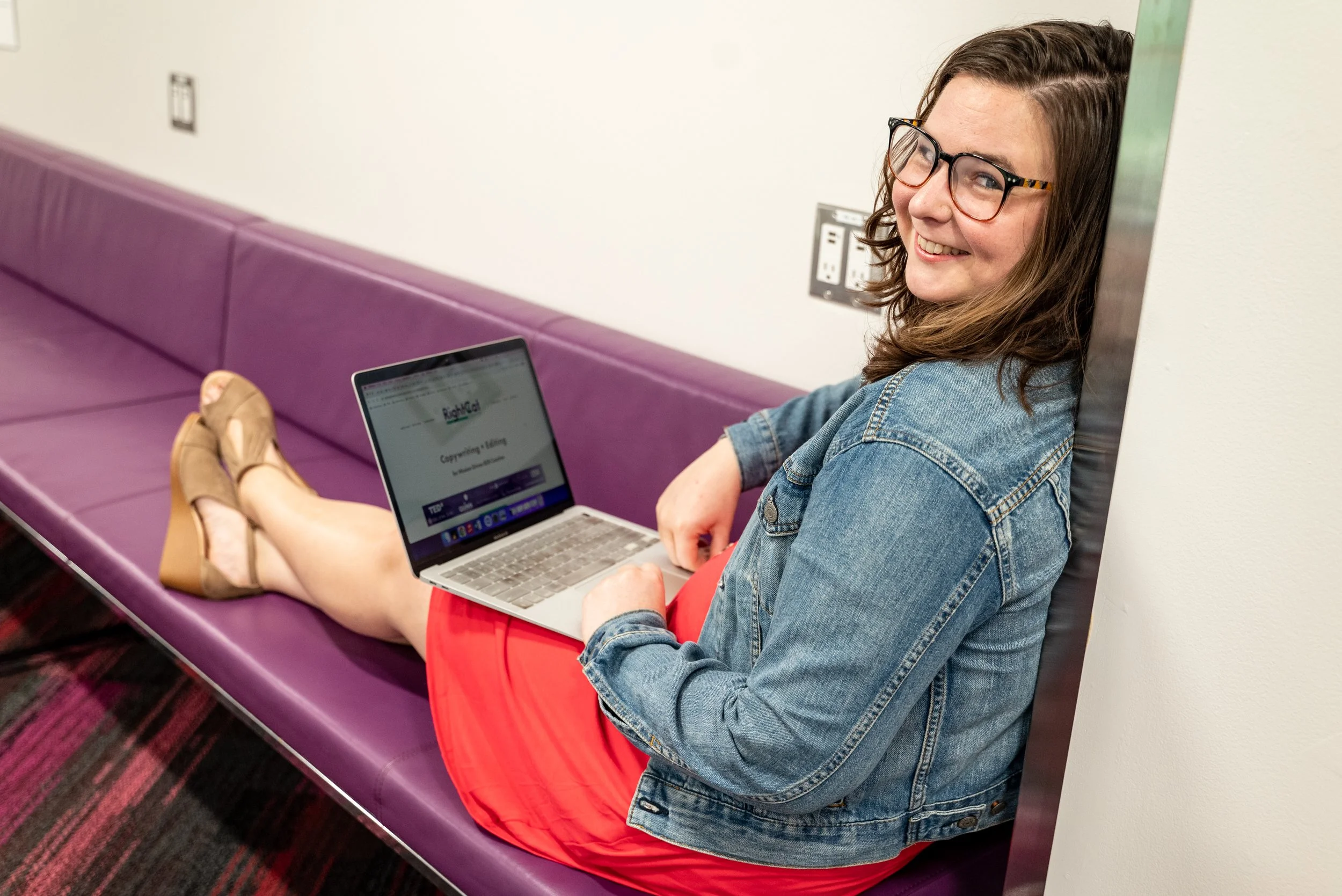 Copywriter + Keynote Coach Cathlyn Melvin leans back on a purple bench with her laptop open on her lap, smiling at the camera