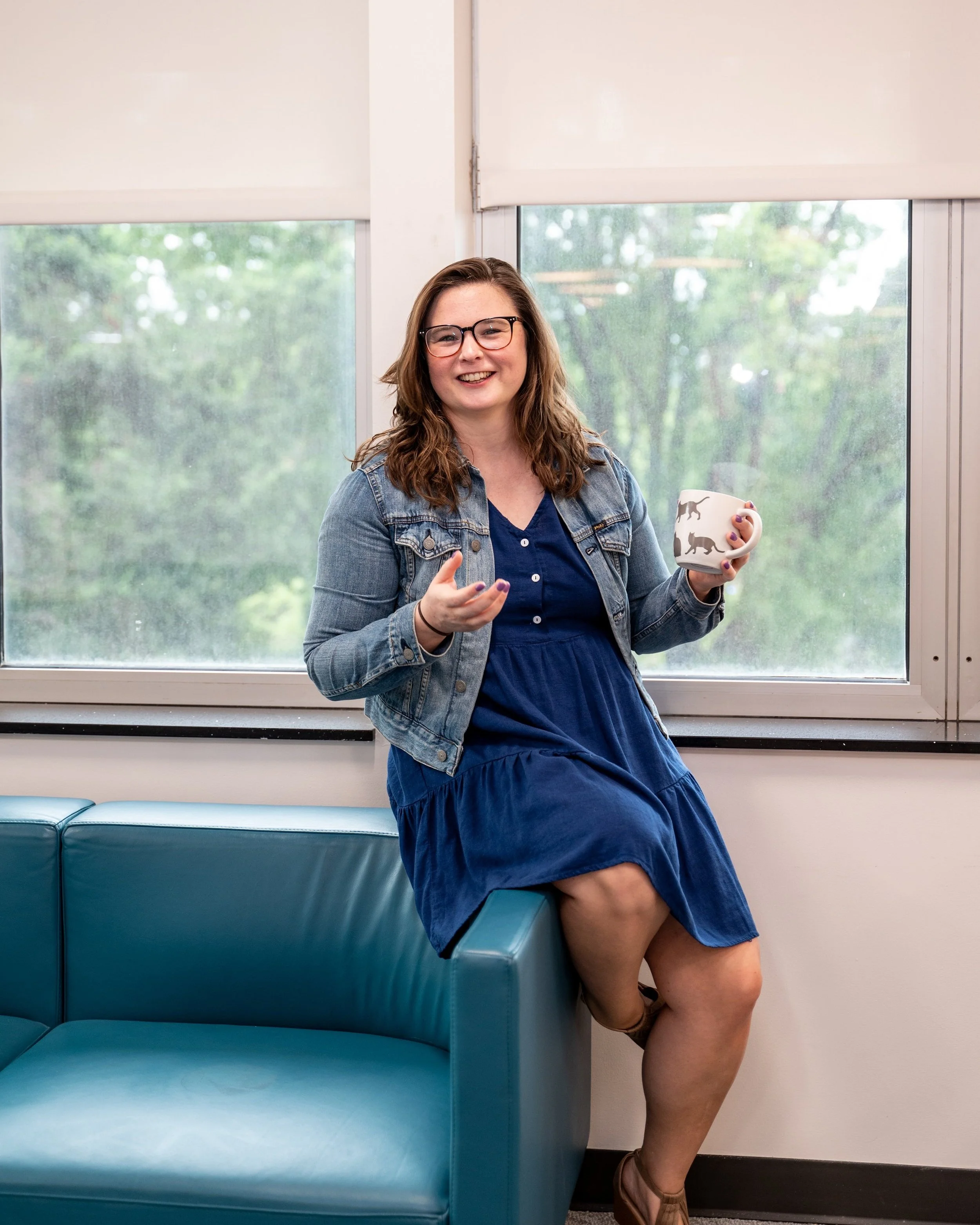 Cathlyn Melvin leans against the arm of a blue sofa, holding a mug with grey cats on it in one hand and gesturing with the other.