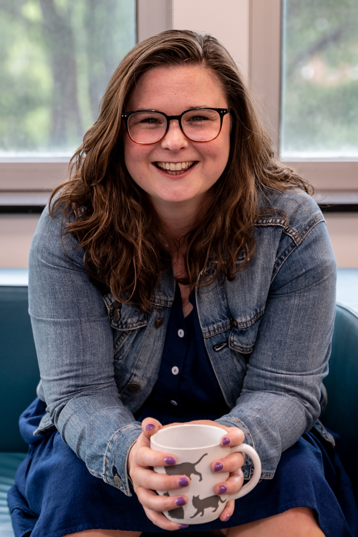 TEDx Coach Cathlyn Melvin, a 30s white woman with brown hair and glasses, leans toward the camera, smiling and holding a large, white coffee mug printed with grey cat silhouettes