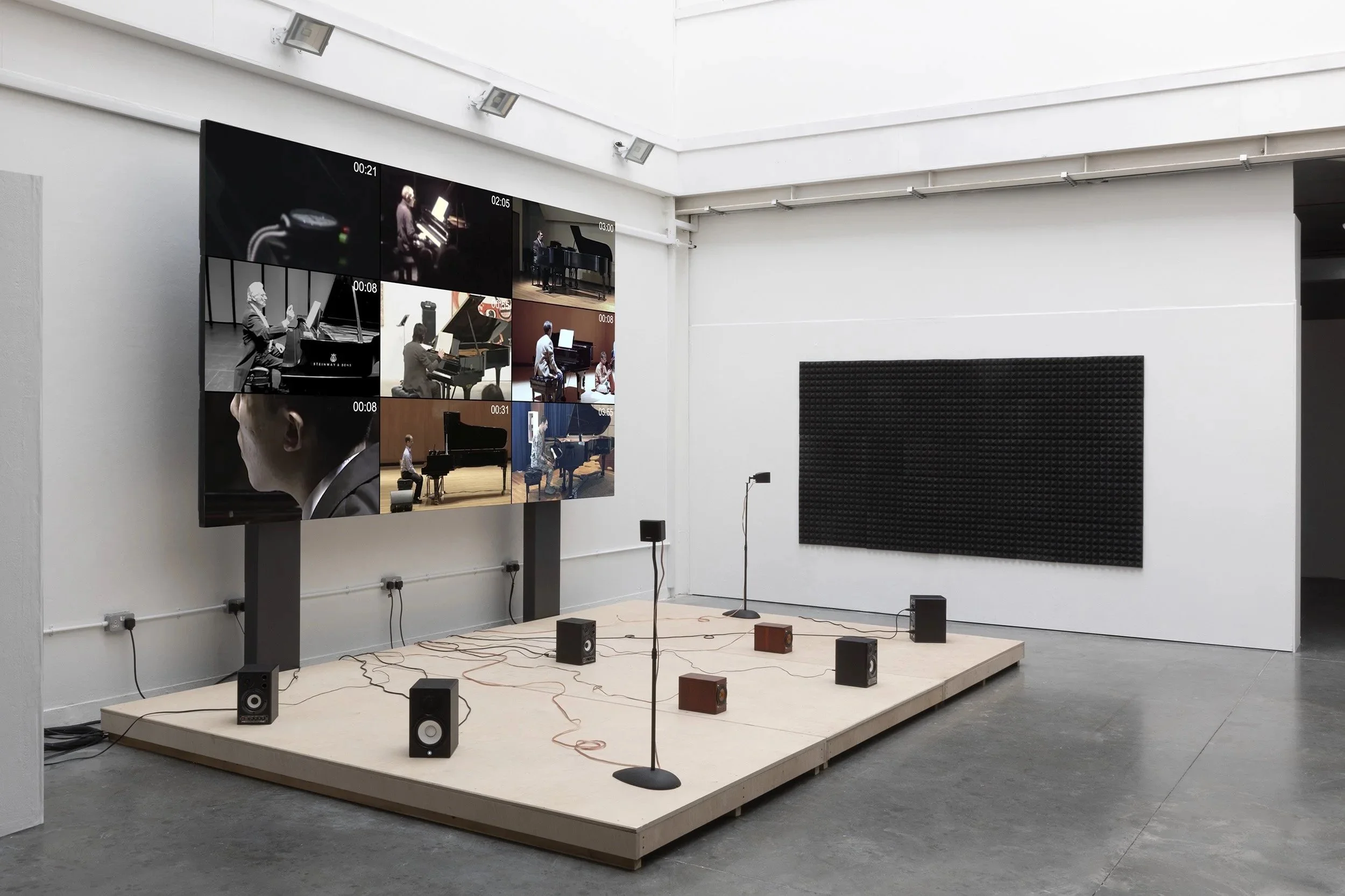 Gallery exhibition, angle view of nine monitor screen plays simultaneous videos of pianist performing at the composition 4'33'' by John Cage. A wooden stage holds nine audio speakers each channeling the sound of silence from one of the performances.