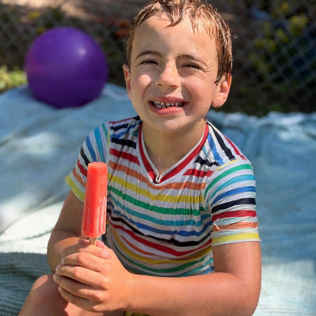 A child smiling in the sun holding a popsicle.
