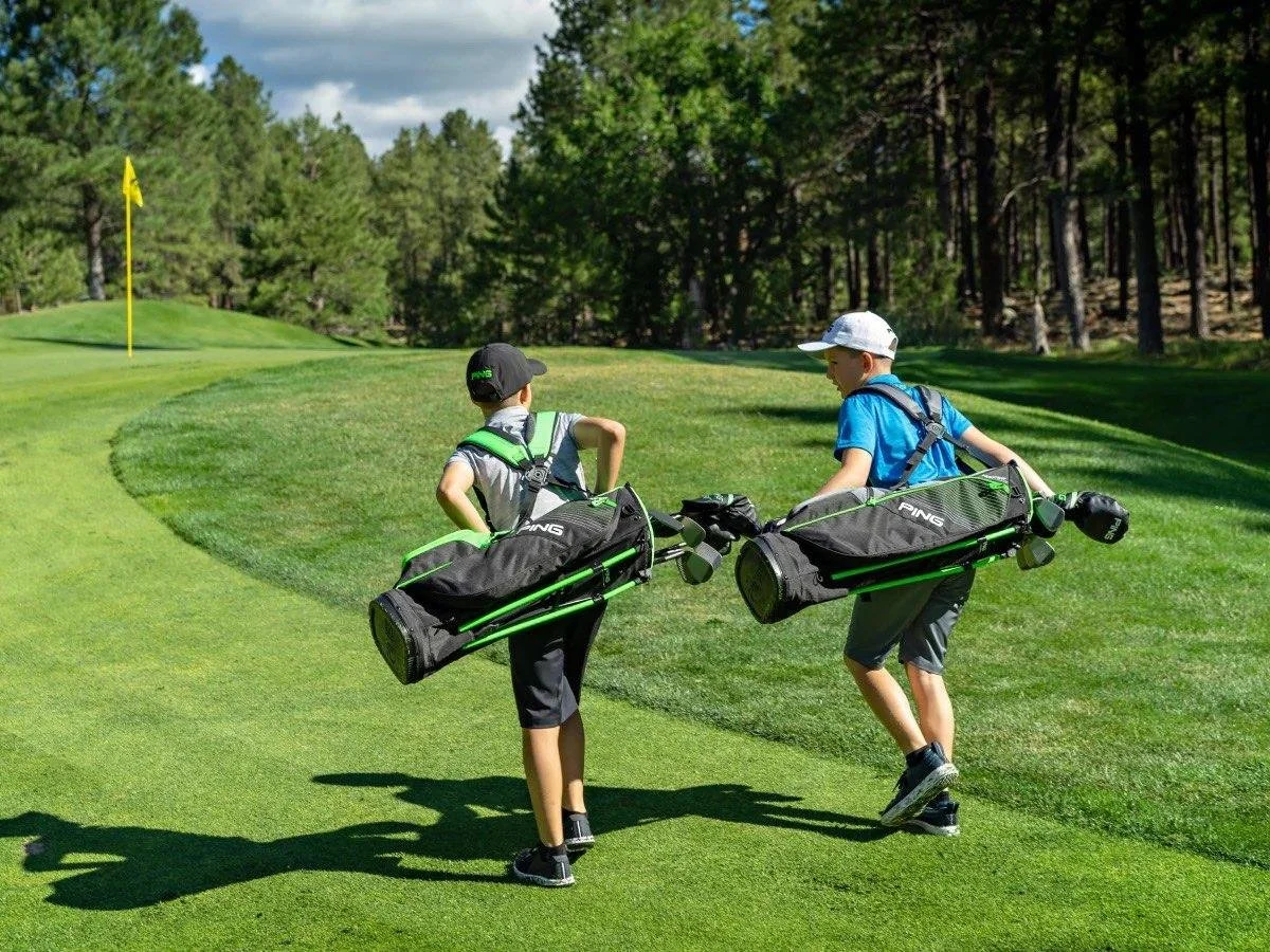 Two boys walking on a golf course, carrying golf bags, towards the green with a flagstick, surrounded by trees.