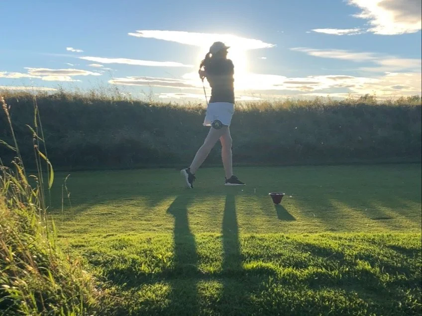 Person playing golf on a grassy course during sunset, holding a golf club, with long shadows cast on the ground.