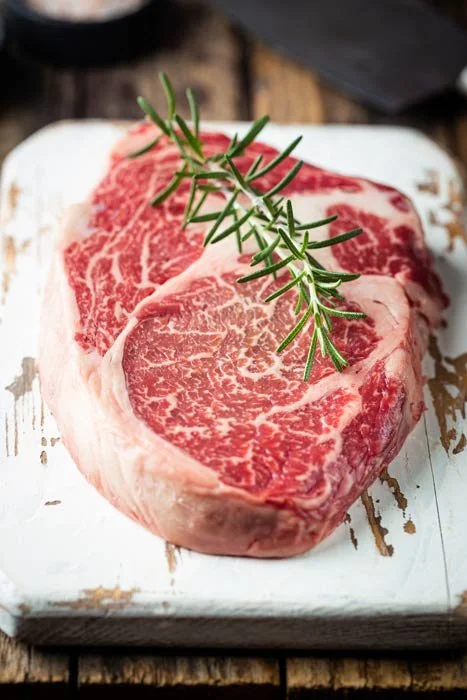 Raw marbled steak with a sprig of rosemary on top, placed on a white cutting board.