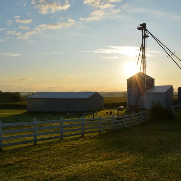 A rural farm scene at sunset featuring a large barn, a grain silo, and farm equipment with a white fence in the foreground.