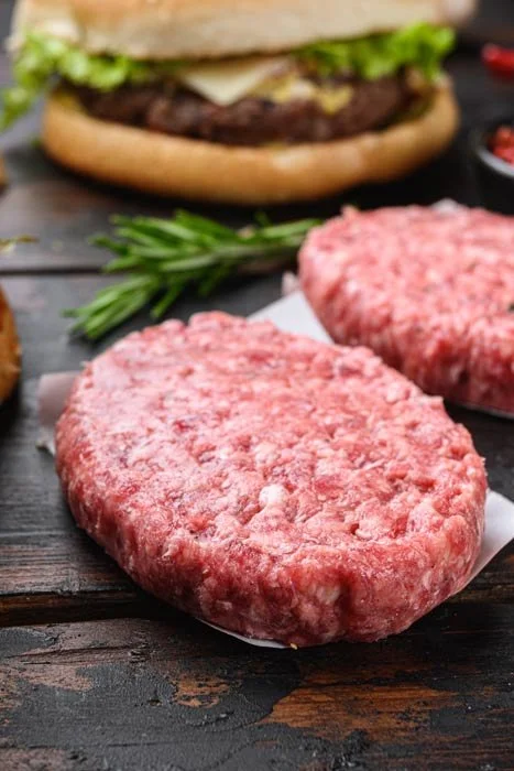 Uncooked ground beef patties on a piece of parchment paper, with a sprig of rosemary and a burger in the background.