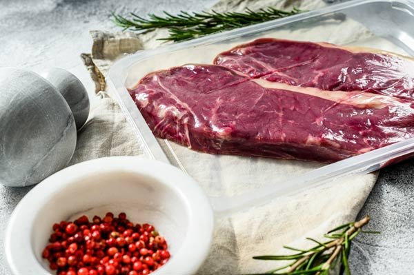 Raw beef steak on a baking sheet with rosemary, pink peppercorns in a bowl, and a gray salt and pepper shaker set.