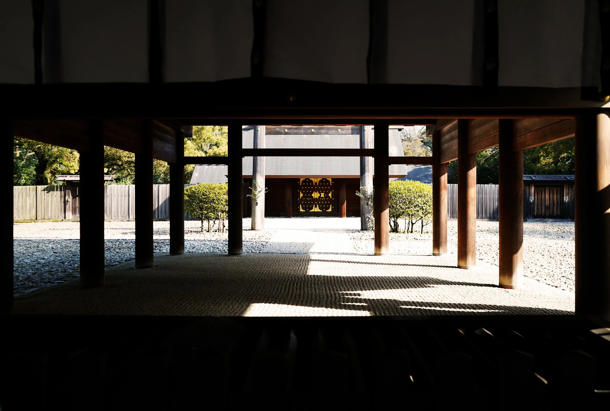 Inside view of a traditional Japanese wooden shrine, showing wooden pillars, a gravel courtyard, and trees outside, with sunlight casting shadows.