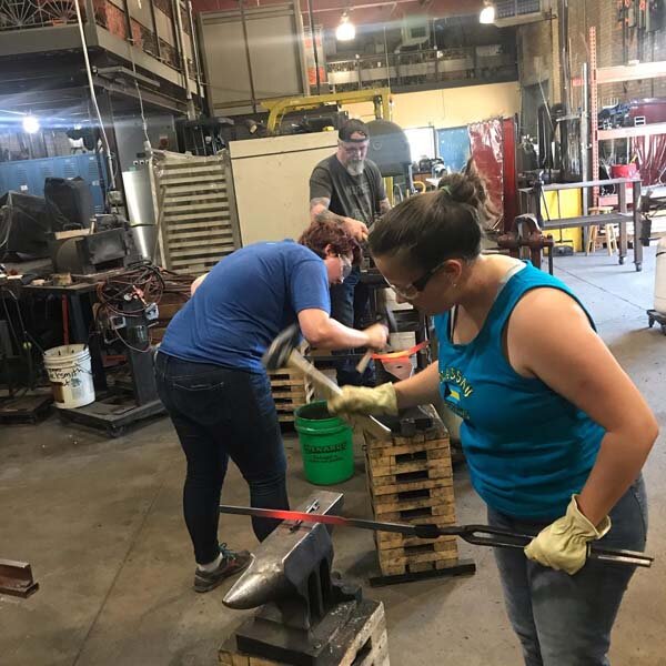  Photo of blacksmithing students swinging hammers. 