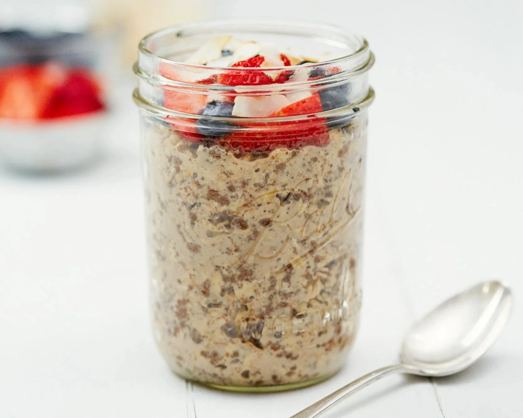 overnight oats in a small jar topped with strawberries and blueberries. a spoon is resting on the table next to the jar.