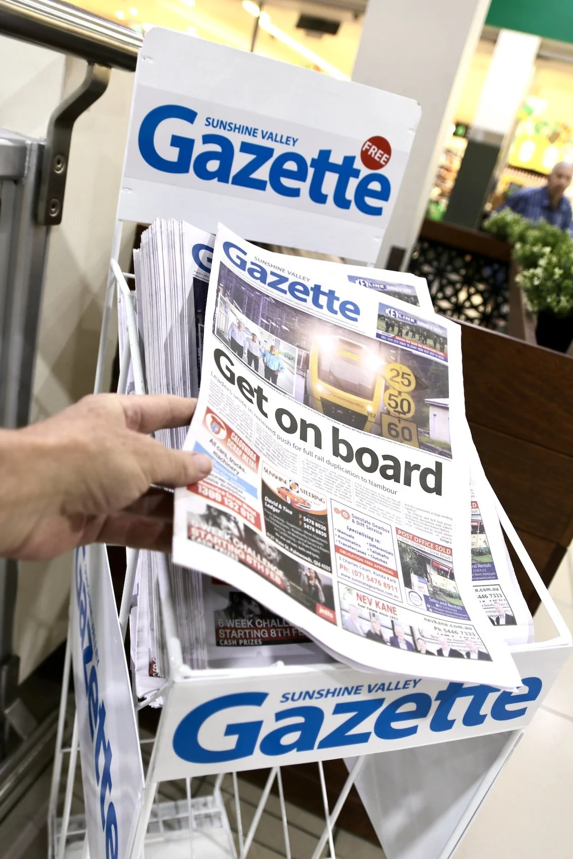A person holding a copy of the Sunshine Valley Gazette newspaper at a newspaper stand in a supermarket, with the headline 'Get on board' and an image of a yellow train.