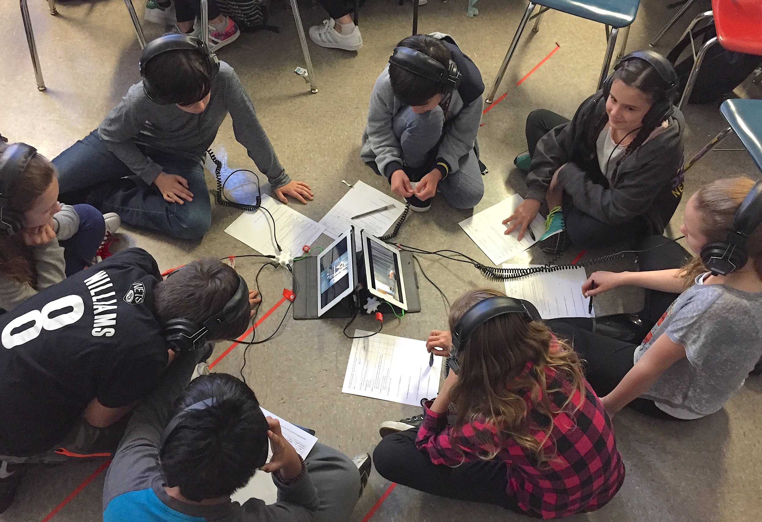  a group of students sit on the floor, wearing headphones, and viewing performances on iPads, while they assess their performances. 