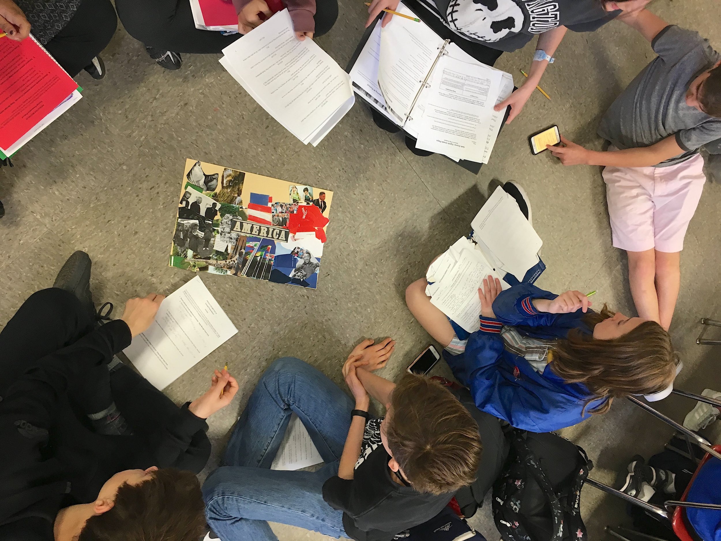  students sit on the floor in a circle reviewing a theatre concept collage 
