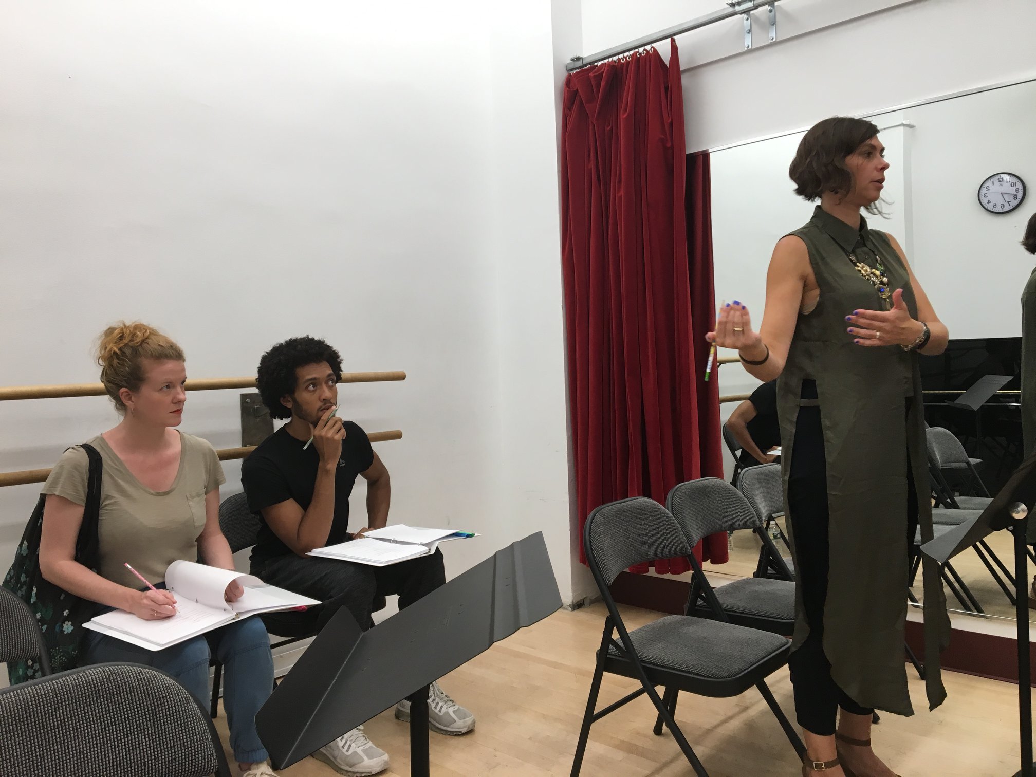  a woman (Aliza) in a long green top stands giving direction to two actors sitting in chairs with scripts on their laps 