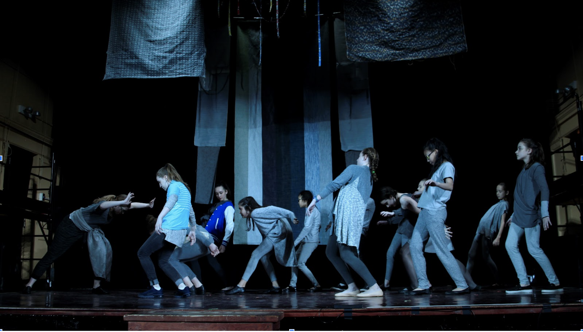  a cool-lit stage with hanging fabric panels and an ensemble of performers in grey in various states of dance 