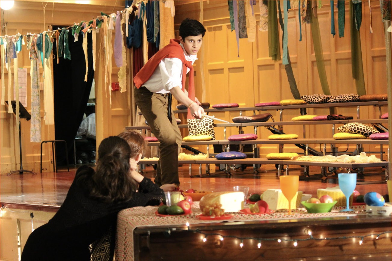 young actor with an orange sweater around their shoulders stands in front of choral bleachers on a state, points a sword towards the camera, disrupting a picnic being held atop a piano. he is surrounded by hanging strips of green fabric 
