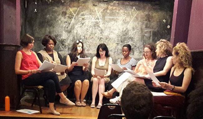  a group of woman sit in chairs on a small stage with scripts on their laps reading a play in front of an audience 